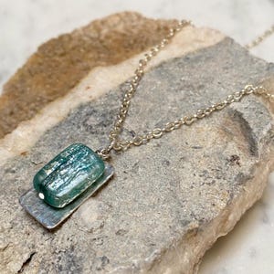 May include: A silver necklace featuring a rectangular, teal-colored pendant atop a hammered silver base. The pendant has a textured appearance, and the chain is delicate. The necklace is displayed on a gray stone surface.