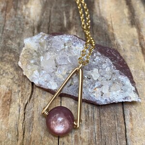 May include: A gold-toned metal necklace featuring a triangular pendant frame that holds a single, smooth, round, mauve-colored gemstone bead. The pendant is suspended from a delicate gold-toned chain, creating a simple and elegant jewelry piece.