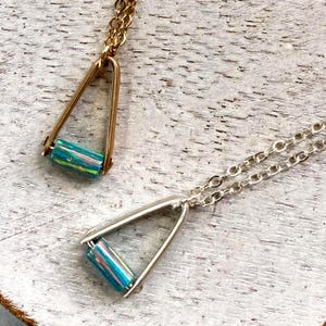May include: Two necklaces featuring triangular metal frames suspended from delicate chains. One necklace has a gold-toned frame and chain, while the other has a silver-toned frame and chain. Each frame holds a cylindrical bead with iridescent blue-green hues.
