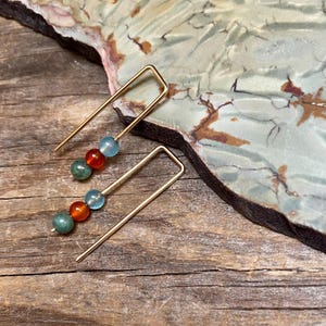 May include: A pair of gold-colored, linear earrings. Each earring features a straight, thin post that curves at the top. The posts are adorned with a series of colorful beads: green, orange, and light blue. The earrings rest on a wooden surface.
