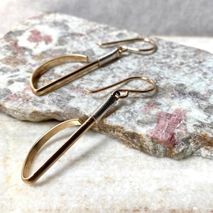 May include: A pair of gold-toned dangle earrings featuring a semi-circular arch design. Each earring has a thin, curved metal bar attached to a straight post, accented with a silver-toned wire wrapping detail near the top hook for suspension.
