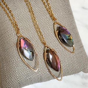 May include: Three gold-colored necklaces with oval-shaped pendants. Each pendant features a colorful, iridescent gemstone. The gemstones display hues of purple, pink, and blue. The necklaces are displayed on a textured, light gray surface.