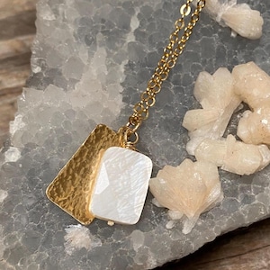 May include: A gold-colored necklace with a rectangular, textured pendant and a white, faceted stone. The necklace has a delicate chain and is displayed on a gray, textured surface with white crystal formations.