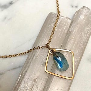 May include: A gold-colored necklace featuring a square pendant with a blue and green gemstone. The gemstone is suspended within the square frame, and the necklace has a delicate chain.