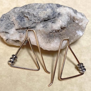 May include: A pair of gold-toned wire earrings shaped as open triangles. Each earring features three small, faceted, dark metallic beads threaded onto one side of the wire frame. The earrings are constructed from a single continuous piece of wire.