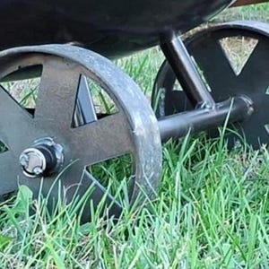 May include: Close-up of a black metal smoker with two large wheels. The wheels have a spoke design and are set in grass.