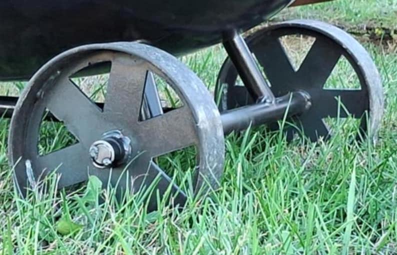 May include: Close-up of a black metal smoker with two large wheels. The wheels have a spoke design and are set in grass.