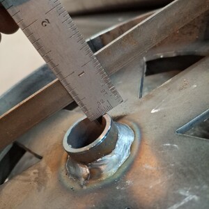 May include: Close-up of a metal pipe with a ruler measuring the diameter of the pipe. The pipe is welded to a metal plate with a star-shaped cutout.