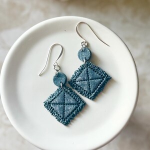 May include: A pair of dangle earrings featuring a small circular stud connected to a larger diamond-shaped pendant. Both components are made of a dark blue textured fabric with visible cross-stitching and frayed edges, suspended by silver hooks.