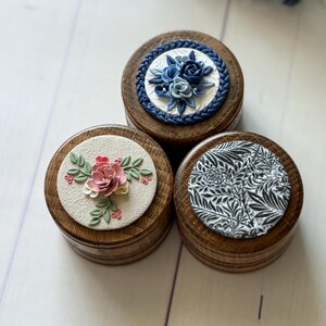 May include: Three round wooden boxes with different floral designs on the lids. The lids are decorated with blue, pink, and black flowers and leaves. The boxes are perfect for storing small items like jewelry or trinkets.