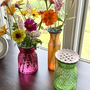 May include: Three glass vases holding colorful wildflowers. One vase is bright pink with a diamond-cut texture, one is a tall frosted orange cylinder, and one is light green with a diamond-cut texture and a white floral-patterned lid.