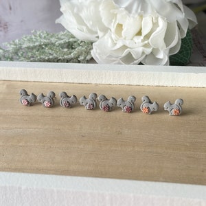 May include: Gray squirrel stud earrings with pink and orange flower accents. The earrings are arranged on a light brown wooden surface, with a white floral arrangement in the background. These earrings are a cute accessory.