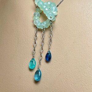 May include: A silver chain necklace with a light green gemstone pendant. The pendant is made of multiple beads and has three silver chains with blue gemstone drops hanging from it.