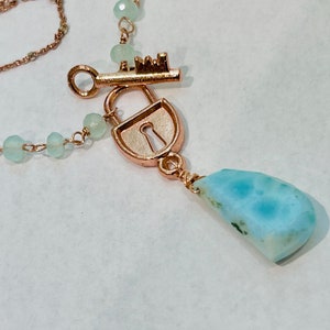 May include: A delicate necklace with a rose gold chain and a pendant featuring a lock and key charm, a light blue gemstone, and light green beads.