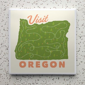 May include: A white square coaster with a green outline of the state of Oregon, with a green and white dotted line representing a road trip route. The text "Visit Oregon" is in orange letters.