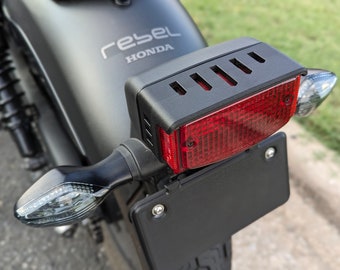 May include: Close-up of a motorcycle's rear section, featuring a black taillight assembly with a red lens and a license plate holder. The motorcycle's body is matte gray, with the word "rebel" and the Honda logo visible.