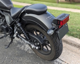 Könnte beinhalten: Ein mattschwarzes Motorrad mit schwarzem Sitz, Heckfender und Reifen. Das Heck des Motorrads hat einen Kennzeichenhalter, ein rotes Rücklicht und einen transparenten Blinker. Das Wort "rebel" ist auf dem Kotflügel zu sehen.