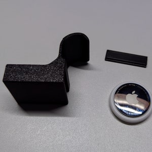 May include: A black 3D-printed holder, a small black rectangular piece, and a white and silver AirTag with the Apple logo. The holder has a textured surface and a curved top. The items are on a light grey surface.