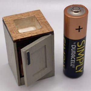 May include: A small wooden cabinet with a light grey door and a brown wooden top. A Duracell Simply AA battery is next to the cabinet. The cabinet is a miniature item.