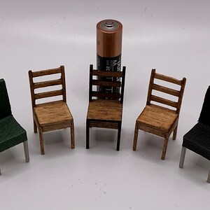 May include: Five miniature chairs in various styles and colours. The chairs include two wooden ladder-back chairs, two chairs with solid backs, and one green chair. A standard AA battery is placed in the centre.