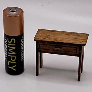 May include: A small wooden table with a drawer, next to a Duracell battery. The table is brown with a rectangular top and four legs. The battery is copper and black, with the word "SIMPLY" printed on it. The table is approximately 10 cm long.