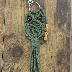 May include: A handmade green macrame owl keychain with brown bead eyes and a silver-tone clasp. The keychain includes orange letter beads spelling "JORDAN" and a long tassel. The owl is made of knotted cord.
