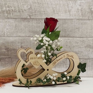 Könnte beinhalten: Holzvase in Form eines Unendlichkeitszeichens mit einer roten Rose und weißen Blumen. Die Vase ist mit dem Datum "29.07.2023" und den Namen "Katharina & Günther" graviert.
