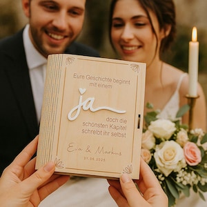 May include: A wooden wedding keepsake box with the words "Eure Geschichte beginnt mit einem ja doch die schönsten Kapitel schreibt ihr selbst Eva & Markus 31.05.2025" engraved on the front. The box is held by a person in front of a couple.