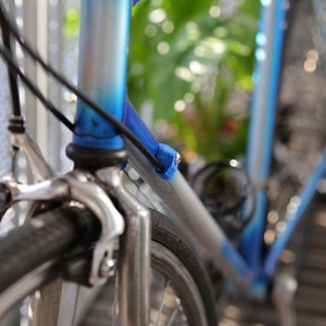 May include: Close-up of a blue and silver bicycle frame with a black cable running along the frame. The front wheel is in focus and has a black tyre with a white rim. The bicycle is parked on a wooden surface.