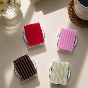 May include: Four rectangular compact mirrors with striped patterns in red, pink, black, and yellow. Each mirror features a clear acrylic casing and is positioned on a circular base. The designs include vertical stripes of varying widths and colour combinations.