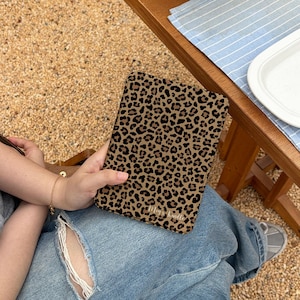 Peut inclure: Une housse de tablette rectangulaire avec un imprimé léopard marron et noir. L'étui est tenu sur les genoux d'une personne, avec un jean bleu et une table en bois en arrière-plan. Le texte "Mays Kindle" est visible sur l'étui.