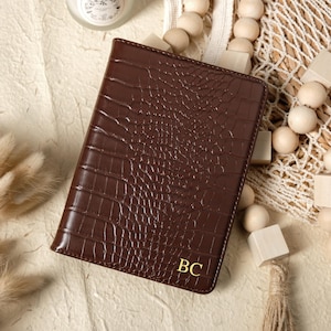 May include: A brown leather passport holder with a crocodile skin texture. The initials "BC" are embossed in gold on the lower right corner. The passport holder is on a neutral-toned surface, with decorative wooden beads and a candle in the background.
