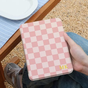 May include: A pink and white checkered tablet case with the initials "MK" in gold. The case is held in a person's lap, with a wooden table and a blue and white striped placemat in the background.