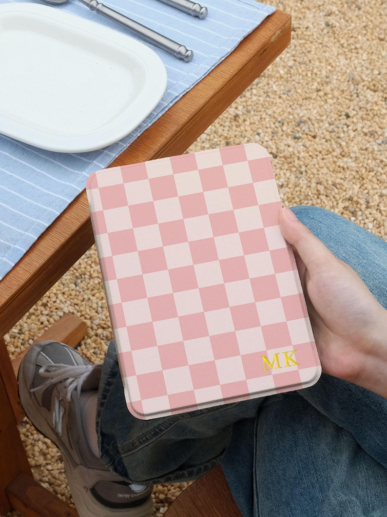 May include: A pink and white checkered tablet case with the initials "MK" in gold. The case is held in a person's lap, with a wooden table and a blue and white striped placemat in the background.