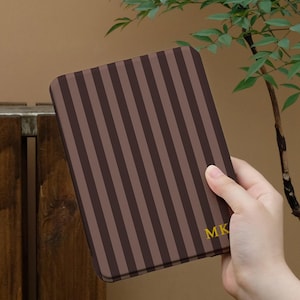 May include: A rectangular e-reader case featuring a vertical striped pattern in alternating shades of dark brown and light brown. The bottom right corner of the case displays the letters MK printed in a bold, yellow, sans-serif font.