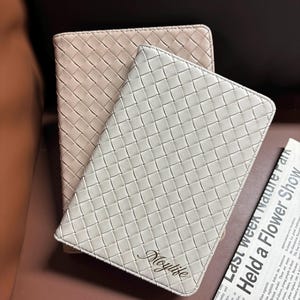 Puede incluir: Dos cuadernos de cuero trenzado, uno beige y otro blanco, con la palabra "Moylife" grabada en el cuaderno blanco.