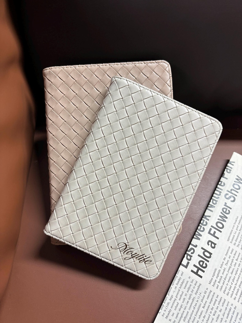 Puede incluir: Dos cuadernos de cuero trenzado, uno beige y otro blanco, con la palabra "Moylife" grabada en el cuaderno blanco.
