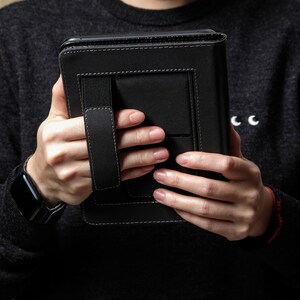 May include: Black leather tablet case with white stitching and a hand strap. The case has a rectangular shape and a secure closure. The person is wearing a black sweater and a smartwatch.