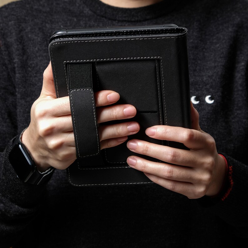 May include: Black leather tablet case with white stitching and a hand strap. The case has a rectangular shape and a secure closure. The person is wearing a black sweater and a smartwatch.