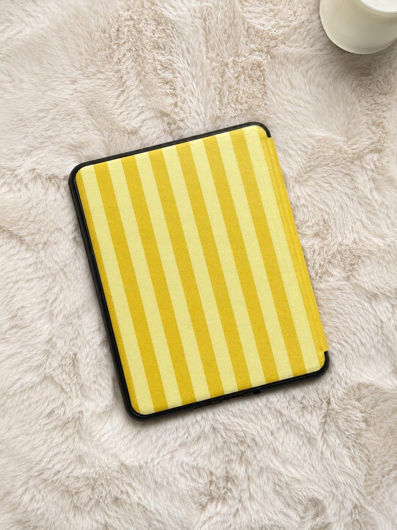 May include: A rectangular e-reader case featuring a vertical striped pattern in alternating shades of bright yellow and pale yellow. The case has a solid black frame surrounding the striped front panel, which is oriented vertically in the center.
