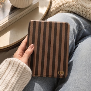 May include: A rectangular e-reader case featuring a vertical striped pattern in alternating shades of dark brown and light brown. The bottom right corner of the case displays the gold-colored embossed initials OB in a serif font.