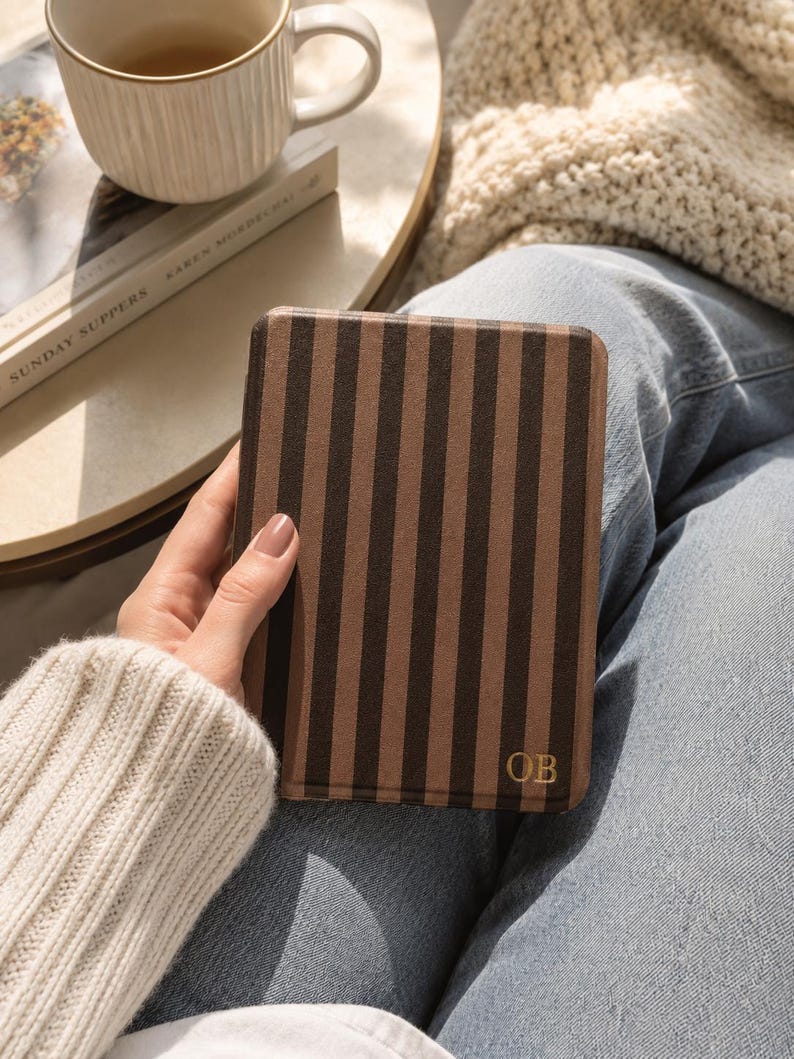 May include: A rectangular e-reader case featuring a vertical striped pattern in alternating shades of dark brown and light brown. The bottom right corner of the case displays the gold-colored embossed initials OB in a serif font.