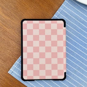 May include: A black-framed tablet case featuring a pink and white checkered pattern. The case is resting on a blue and white striped cloth, with a wooden surface in the background.