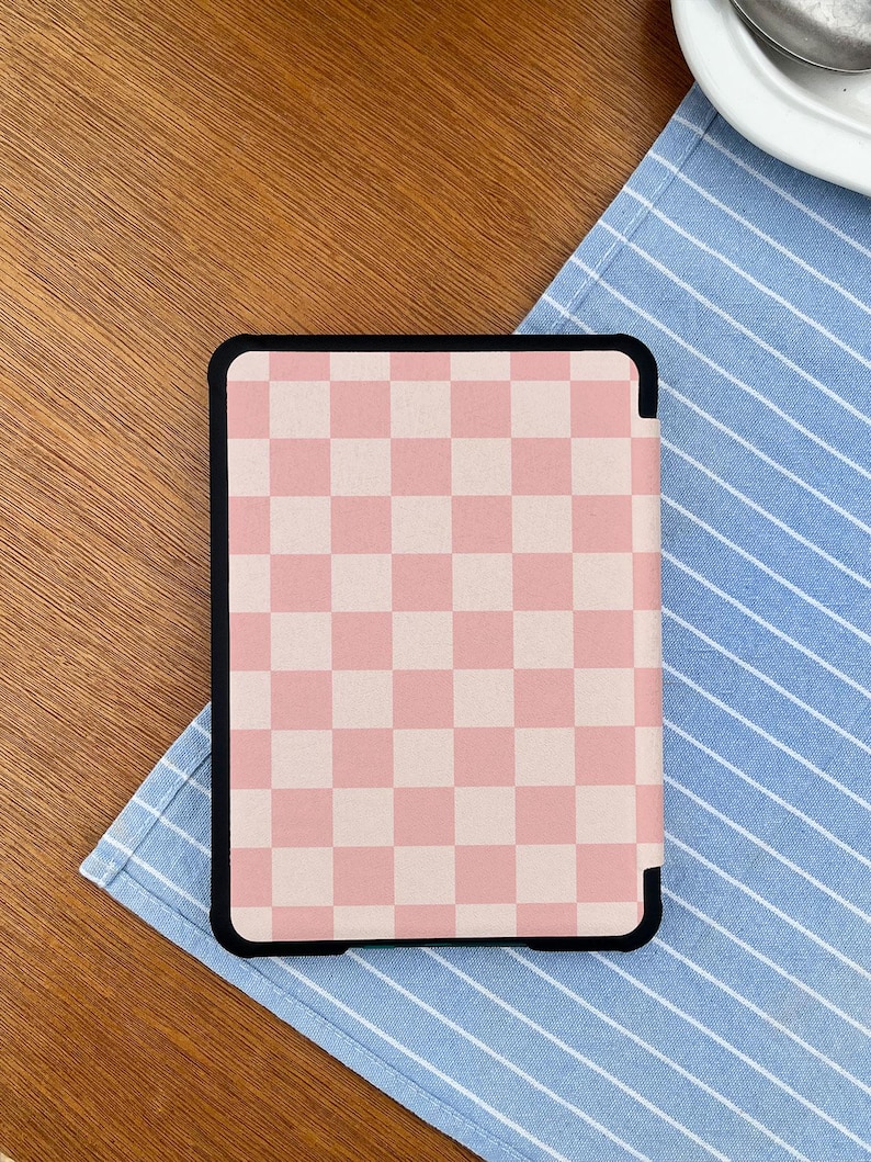 May include: A black-framed tablet case featuring a pink and white checkered pattern. The case is resting on a blue and white striped cloth, with a wooden surface in the background.