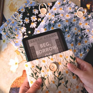 以下が含まれることがあります： クリーム、黒、水色の刺繍入り電子書籍リーダーケース。デイジー柄で装飾されています。各ケースにはボタン留めがあり、Sarah Adamsの「Beg, Borrow, Or」というテキストが表示された電子書籍リーダーを収納できます。魅力的で実用的なアクセサリーです。