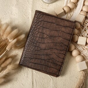 May include: A brown leather passport holder with a crocodile skin texture. The cover is rectangular and closed. The background includes decorative elements such as wooden beads and dried flowers.