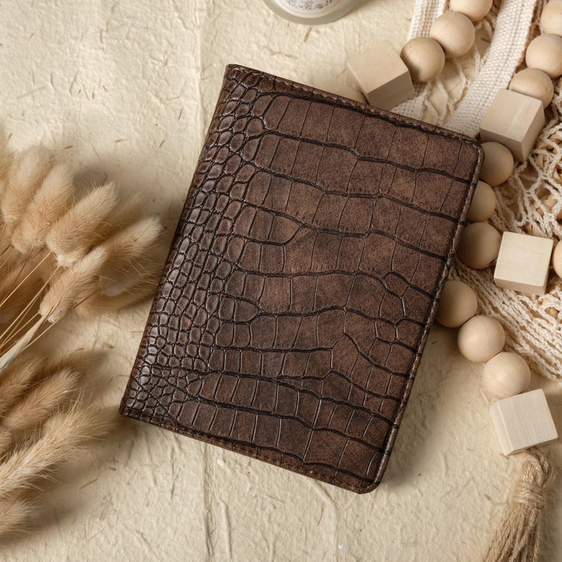 May include: A brown leather passport holder with a crocodile skin texture. The cover is rectangular and closed. The background includes decorative elements such as wooden beads and dried flowers.
