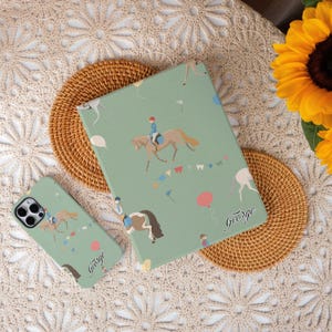 May include: A green phone case and notebook with a horse riding theme. The case features a white horse and rider wearing a blue shirt and the text "George". The notebook has a similar design with a white horse and rider wearing a blue shirt and the text "George".