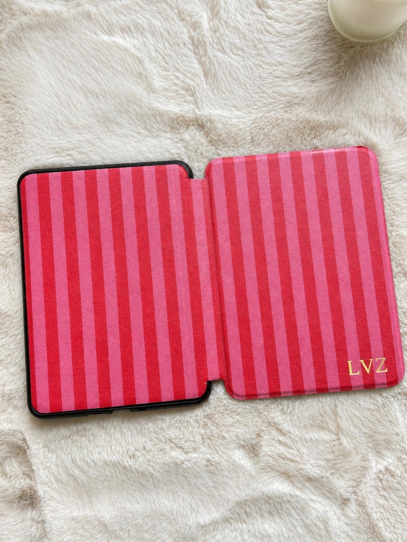 May include: A rectangular e-reader case featuring a vertical striped pattern in alternating shades of bright pink and dark red. The bottom right corner of the case displays the letters LVZ in a gold-colored font. The case is open and flat.