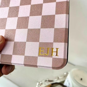 May include: A close-up of a case with a pink and brown checkered pattern. The initials "EJH" are displayed in gold lettering on the lower right corner of the case. The case is held by a hand.